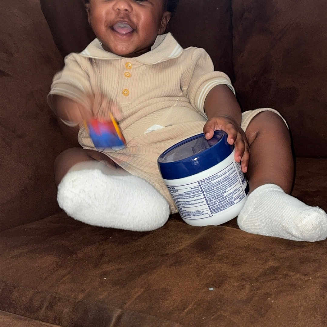 Corday is registered to the contest to win money with this photo: baby, child, smiling, couch, indoor, toy, container, white_socks, happy, person, clothing, furniture, cute, infant, seated, home, cozy, portrait, brown, relaxed