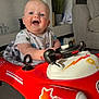 baby, child, toy_car, smiling, indoor, blue_eyes, happy, seat, clothing, car, wheel, living_room, plant, sofa, carpet, head, person, face, play, fun