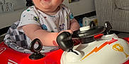 Noah is registered to the contest to win money with this photo: baby, child, toy_car, smiling, indoor, blue_eyes, happy, seat, clothing, car, wheel, living_room, plant, sofa, carpet, head, person, face, play, fun