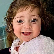 Sophia is registered to the contest to win money with this photo: baby, child, smile, happy, face, portrait, indoor, clothing, quilted_jacket, curly_hair, person, hand, holding, cute, infant, young_child, bright_eyes, expression, warm, soft_lighting