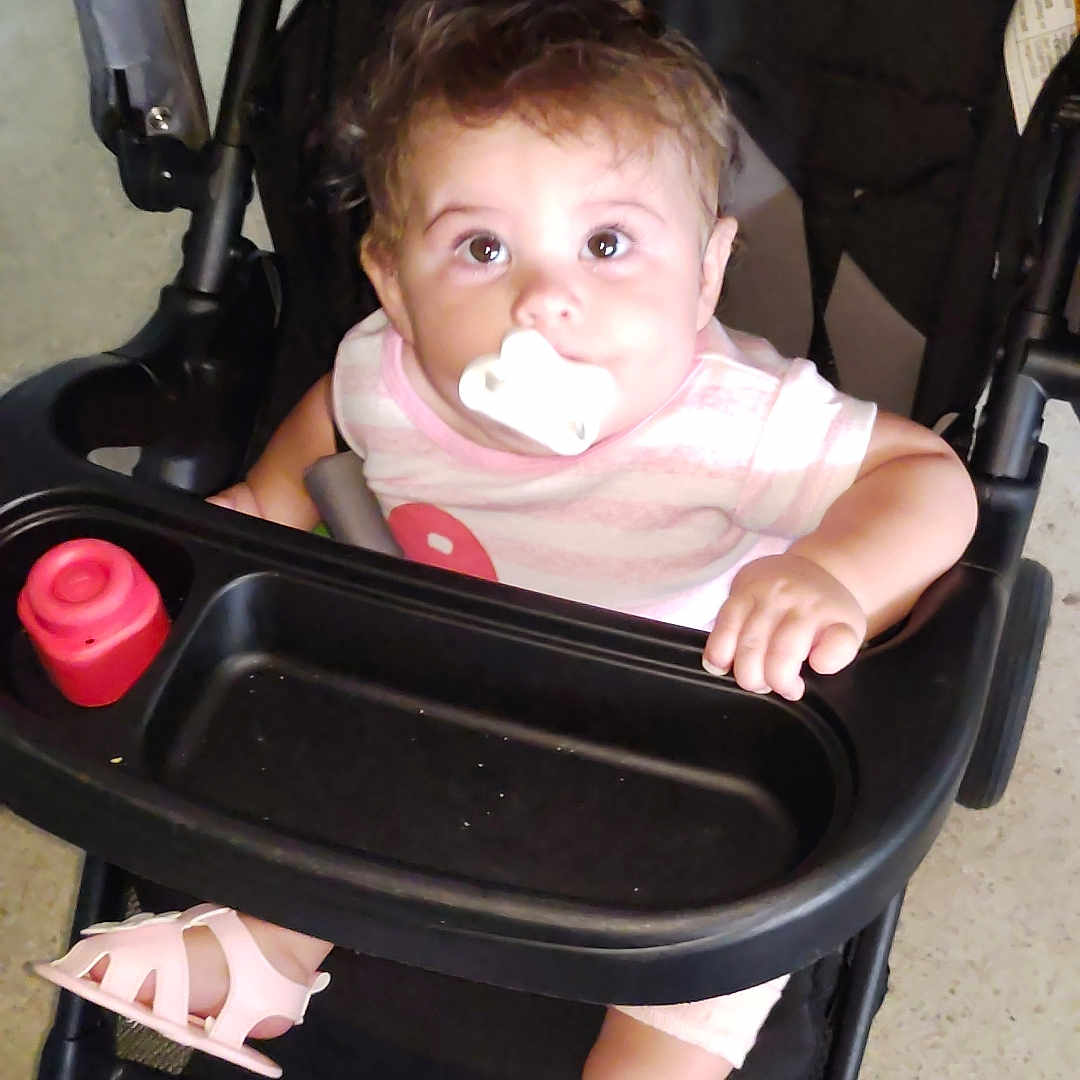 Natalie is registered to the contest to win money with this photo: baby, black_stroller, child, curious, cute, expression, face, floor, footwear, hand, indoor, infant, looking_up, pacifier, pink_sandals, seated, striped_shirt, stroller, toy, young_child