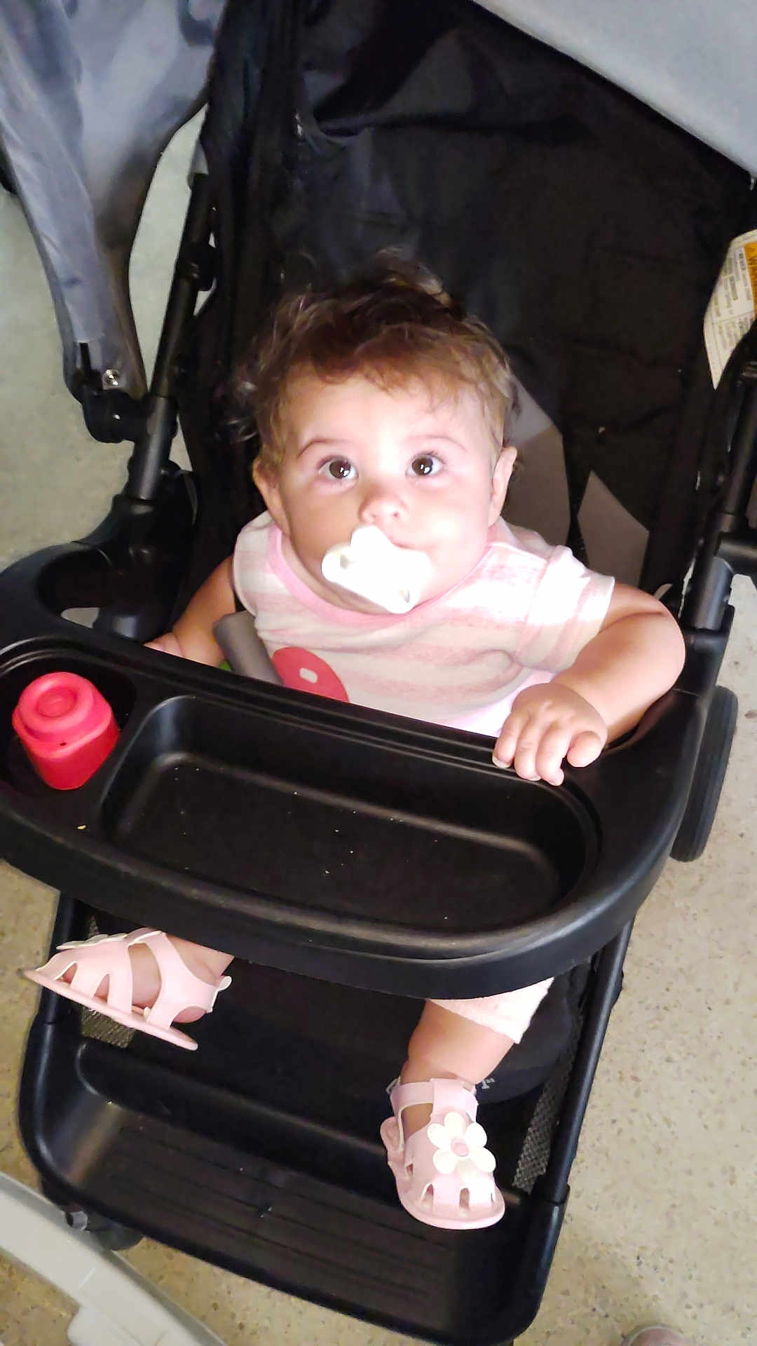 Natalie is registered to the contest to win money with this photo: baby, stroller, pacifier, pink_sandals, striped_shirt, child, indoor, curious, seated, footwear, toy, infant, cute, hand, looking_up, face, young_child, black_stroller, floor, expression