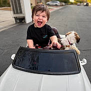 Knoah is registered to the contest to win money with this photo: child, toddler, smiling, toy_car, dog, pet, outdoor, street, suburban, happy, black_shirt, short_hair, daylight, playful, vehicle, steering_wheel, cute, pet_collar, animal, fun