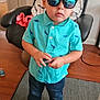 toddler, child, indoor, standing, cap, sunglasses, turquoise_shirt, dinosaur_print, jeans, sneakers, carpet, chair, furniture, hand_gesture, reflection, casual_clothing, young_child, portrait, home_interior, floor