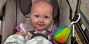 Delilah is registered to the contest to win money with this photo: baby, stroller, blanket, plush_toy, avocado_toy, face, blue_eyes, smile, pink_clothing, beaded_charm, name_charm, seatbelt, child, indoor, shopping_cart, floor_tiles, soft_texture, cute, infant, person