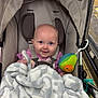 baby, stroller, blanket, plush_toy, avocado_toy, face, blue_eyes, smile, pink_clothing, beaded_charm, name_charm, seatbelt, child, indoor, shopping_cart, floor_tiles, soft_texture, cute, infant, person