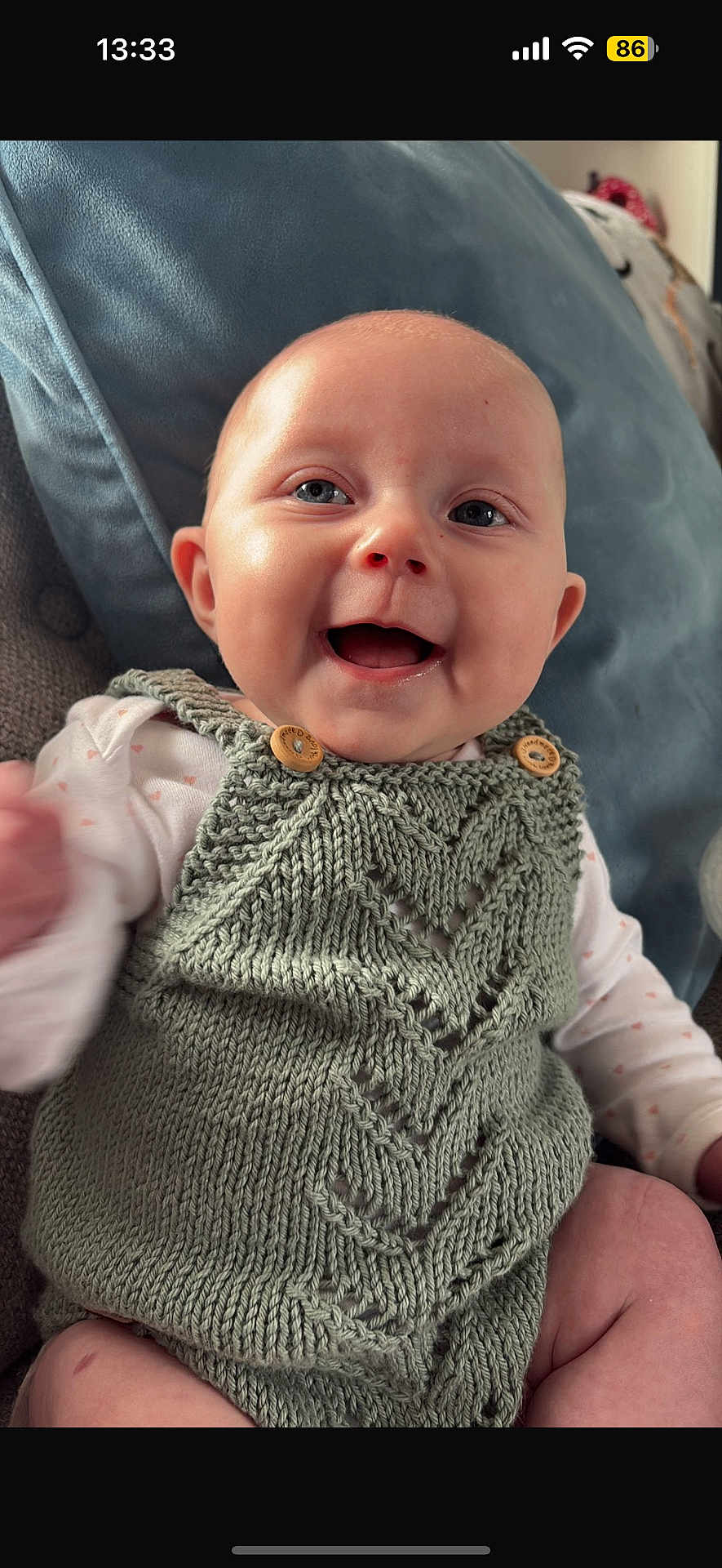 Delilah is registered to the contest to win money with this photo: baby, smiling, infant, knitted_clothing, green_outfit, cushion, indoor, happy, portrait, cute, child, face, skin, person, seated, cozy, handmade, texture, soft, expression