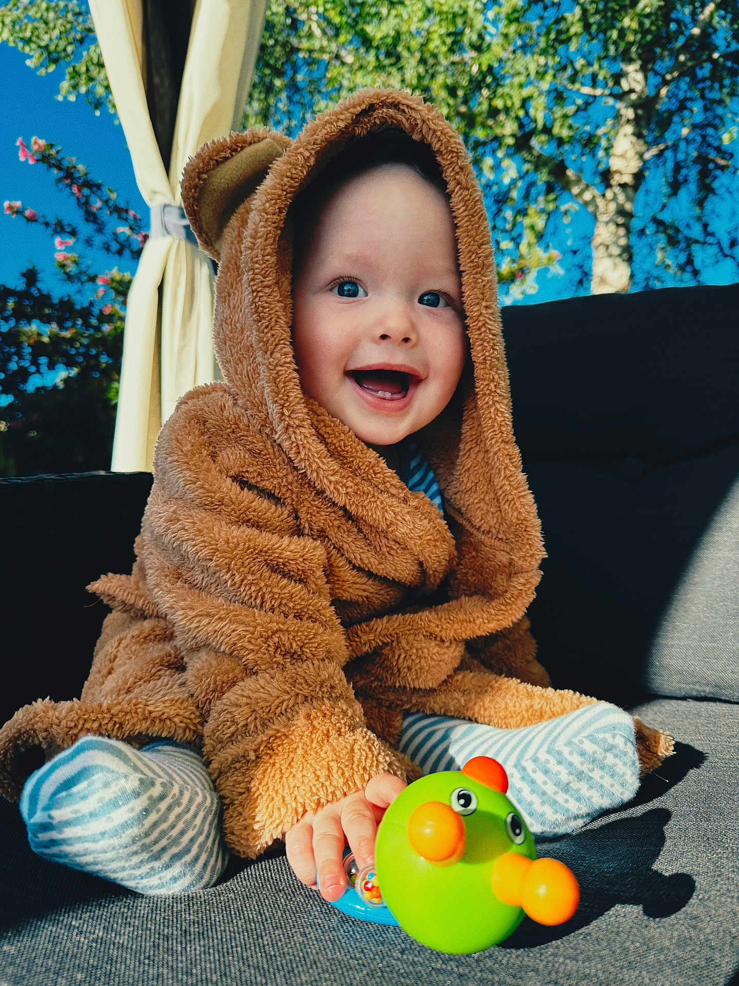 Markas is registered to the contest to win money with this photo: baby, child, toddler, smiling, toy, outdoor, blue_sky, greenery, couch, fuzzy_clothing, hooded_jacket, striped_socks, happy, playful, cute, portrait, sunlight, nature, bright_colors, infant
