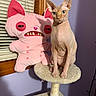 sphynx_cat, hairless_cat, cat_tree, stuffed_toy, pink_toy, window_blinds, purple_wall, wooden_frame, indoor, pet, animal, feline, sitting, portrait, closeup, cute, odd_toy, plush, decor, domestic
