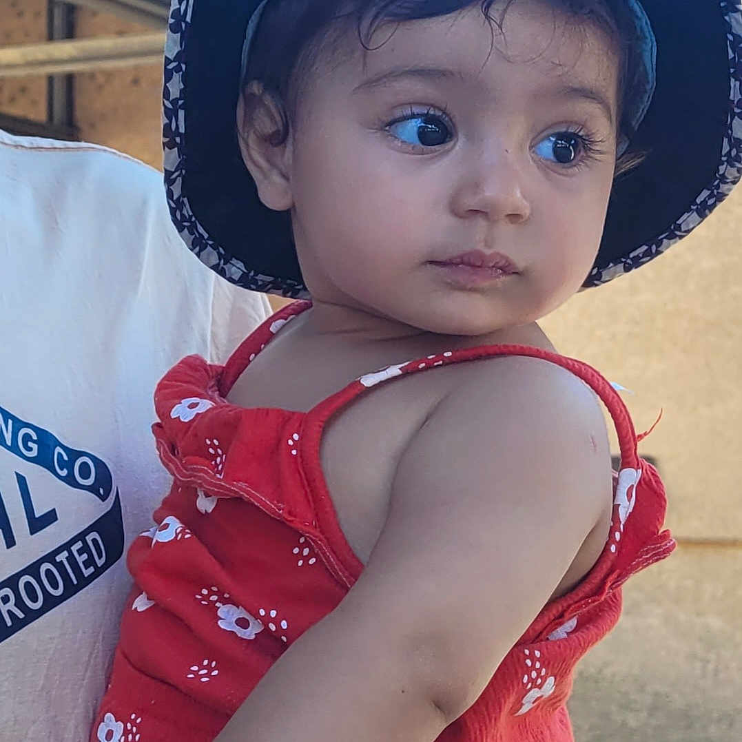 Tasnîm a rejoint le concours — aidez-le/la à gagner de superbes lots ! adult, baby, casual, child, clothing, curious, expression, eyes, face, floral_pattern, hat, holding, outdoor, person, portrait, red_dress, skin, summer, toddler, young_child