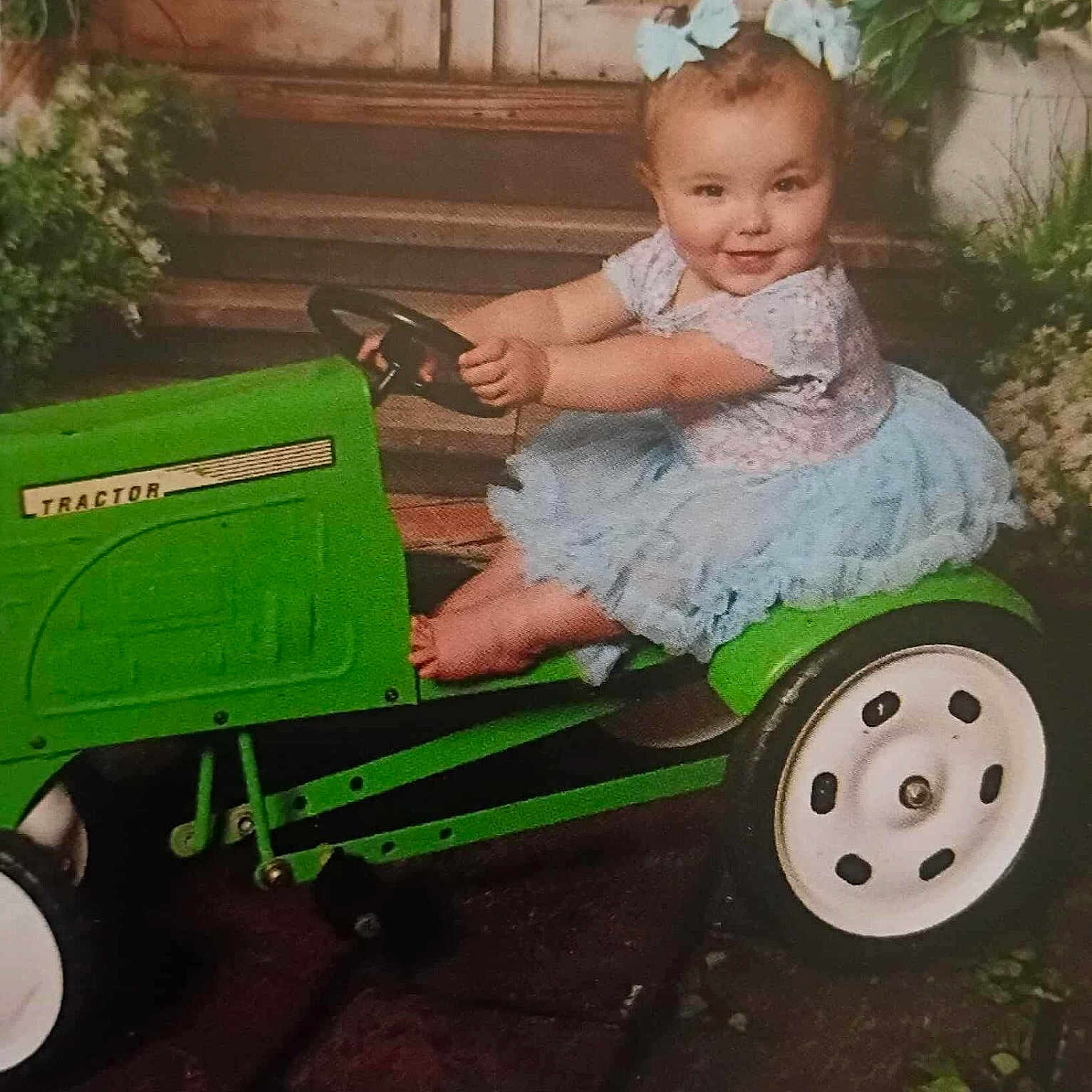 Alyson is registered to the contest to win money with this photo: baby, barn_door, blue_dress, bows, child, cute, feet, fun, girl, green, happy, outdoor, plants, play, portrait, riding, smiling, toy_tractor, vehicle, young