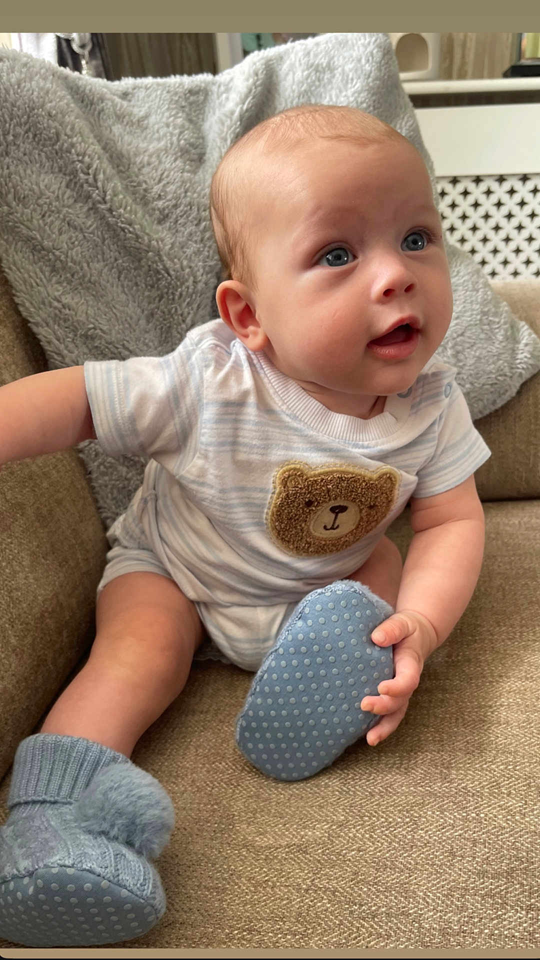 Rueben is registered to the contest to win money with this photo: baby, infant, child, couch, blanket, blue_eyes, onesie, teddy_bear_patch, booties, soft_toys, indoor, portrait, cute, smiling, seated, cozy, furniture, hand, face, expression