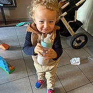 Ewen participe au concours pour gagner de l'argent avec cette photo : toddler, child, baby_bottle, curly_hair, spiderman_shoes, vest, pants, stroller, tile_floor, toy, receipt, indoor, person, footwear, baby, cute, standing, smiling, clothing, playtime