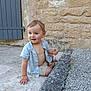 toddler, child, sitting, stone_floor, gravel, blue_shirt, beige_shorts, bare_chest, curious, happy, outdoor, stone_wall, door, hand, feet, person, portrait, young_child, casual_clothing, daylight