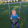 child, toddler, swing, playground, grass, fence, blue_shirt, animal_print_pants, blue_shoes, sunlight, happy, smiling, outdoor, daytime, play, fun, chain, seat, park, nature