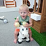 toddler, child, toy, cow, indoor, play_area, carpet, green, orange, sleeveless_shirt, shorts, barefoot, plastic_toy, happy, smiling, blonde_hair, playful, fun, furniture, window