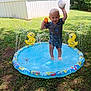 toddler, child, splash_pool, water, grass, yellow_duck, toy, ball, outdoor, summer, play, backyard, sunny, water_spray, smiling, short_sleeves, barefoot, house, greenery, fun
