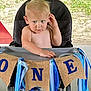 toddler, child, high_chair, banner, birthday, one_year_old, blue_ribbons, outdoor, grass, celebration, blonde_hair, curious_expression, face, hand, seat, person, event, party, skin, young_child