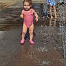 Iyla is registered to the contest to win money with this photo: toddler, child, splash_pad, water, swimsuit, pink_shoes, toy, outdoor, summer, play, water_splash, pavement, sunny, people, bench, shade, trees, relaxing, background, fun
