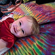 Iyla is registered to the contest to win money with this photo: baby, child, smiling, red_dress, tie_dye_blanket, colorful, indoor, living_room, leather_chair, table, hand, tattoo, face, portrait, happy, candid, person, infant, cozy, casual