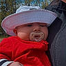 baby, child, pacifier, hat, red_jacket, clothing, adult, glasses, smile, fist, bracelet, outdoor, trees, blue_sky, face, person, holding, portrait, closeup, nature