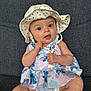 baby, infant, child, hat, polka_dot, dress, floral_pattern, sitting, couch, fabric, blue_eyes, cute, indoors, portrait, young_child, clothing, skin, face, hand, expression