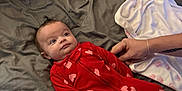 Poppy is registered to the contest to win money with this photo: baby, infant, red_onesie, hearts, bed, blanket, hands, sleepwear, indoors, person, soft_fabric, comfort, cute, lying_down, sleeping_area, portrait, newborn, child, cozy, expression