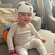 Sophia joined the competition — help win amazing prizes! baby, child, helmet, protective_gear, couch, pajamas, polka_dots, smile, indoor, cozy, cushion, plush_toy, blanket, living_room, window, curtains, furniture, person, sitting, happy
