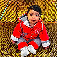 Taqdeer is registered to the contest to win money with this photo: baby, child, red_jacket, gray_pants, socks, golden_mesh, sitting, curious_expression, wet_hair, indoors, bright_colors, cute, portrait, young_child, warm_clothing, face, small_hands, casual_pose, floor_surface, playful