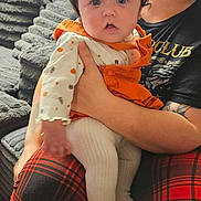 Calliope joined the competition — help win amazing prizes! baby, child, infant, portrait, sitting, held, smiling, wide_eyed, bows, pigtails, orange_outfit, plaid_pants, adult, caregiver, couch, blanket, window, blinds, indoor, home