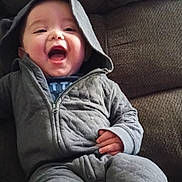 James is registered to the contest to win money with this photo: baby, child, hood, jacket, smile, laughing, couch, seated, cozy, gray_clothing, infant, cute, happy, indoor, person, young_child, fabric, zipper, relaxed, portrait