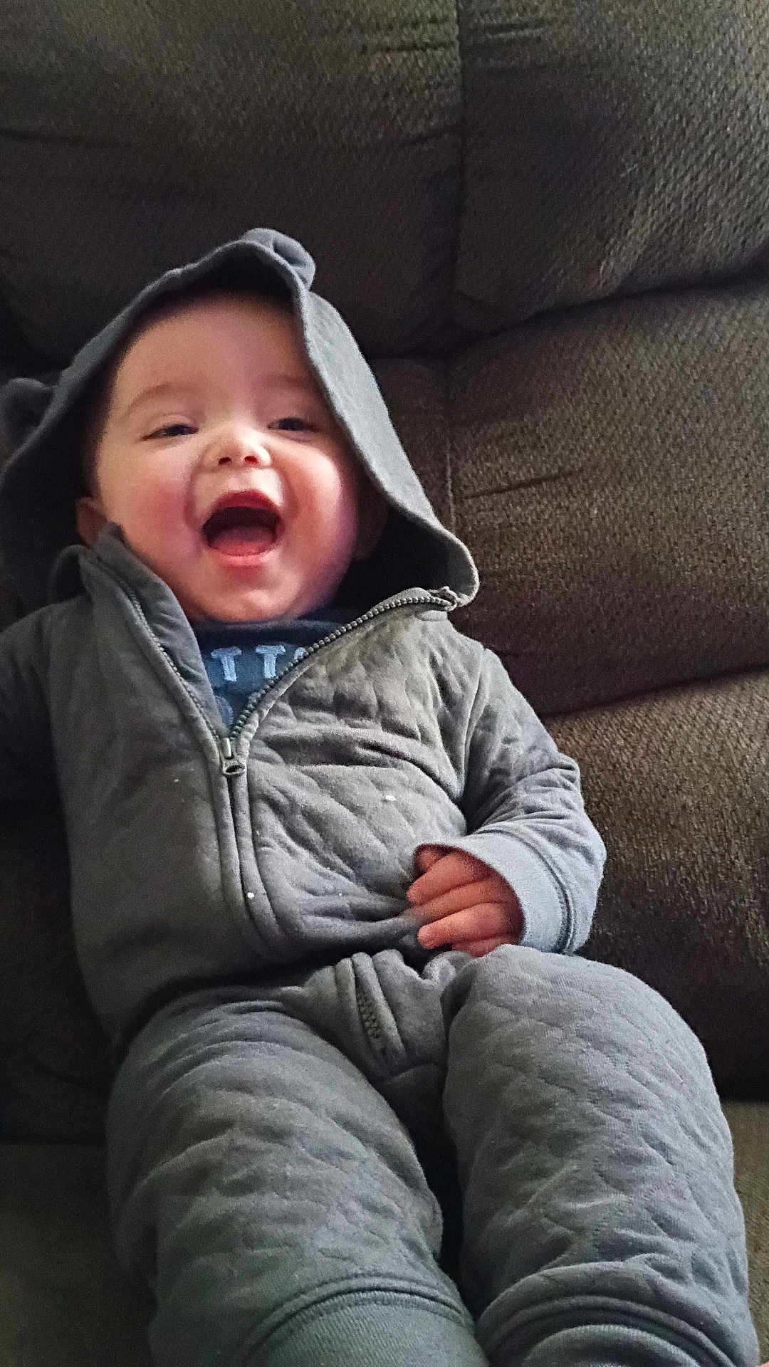 James is registered to the contest to win money with this photo: baby, child, hood, jacket, smile, laughing, couch, seated, cozy, gray_clothing, infant, cute, happy, indoor, person, young_child, fabric, zipper, relaxed, portrait