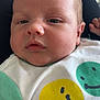 baby, infant, closeup, face, smiling_face, clothing, white_shirt, green_sleeves, yellow_smiley, green_smiley, person, skin, head, expression, cute, newborn, portrait, resting, soft_light, indoors
