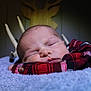 baby, sleeping, plaid_shirt, red_clothing, blue_blanket, soft_texture, wooden_deer_head, antlers, close_up, face, peaceful, indoors, portrait, newborn, child, resting, cozy, warm_lighting, background_blur, cute