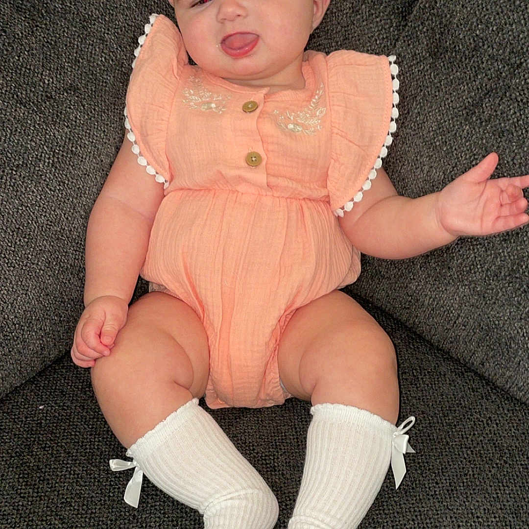 Arizona is registered to the contest to win money with this photo: baby, child, chubby_cheeks, couch, cute, fabric_texture, furniture, happy, headband, indoor, infant, peach_outfit, person, portrait, sitting, smiling, sock, tongue_out, wearing, white_sock