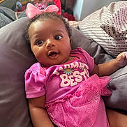 Egypt Ivy Elizabeth is registered to the contest to win money with this photo: baby, child, infant, portrait, indoors, sitting, lying_down, pink_outfit, pink_bow, dress, smiling, wide_eyes, curly_hair, couch, cushions, blanket, toy_storage, home, close_up, fashion