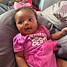 baby, child, infant, portrait, indoors, sitting, lying_down, pink_outfit, pink_bow, dress, smiling, wide_eyes, curly_hair, couch, cushions, blanket, toy_storage, home, close_up, fashion