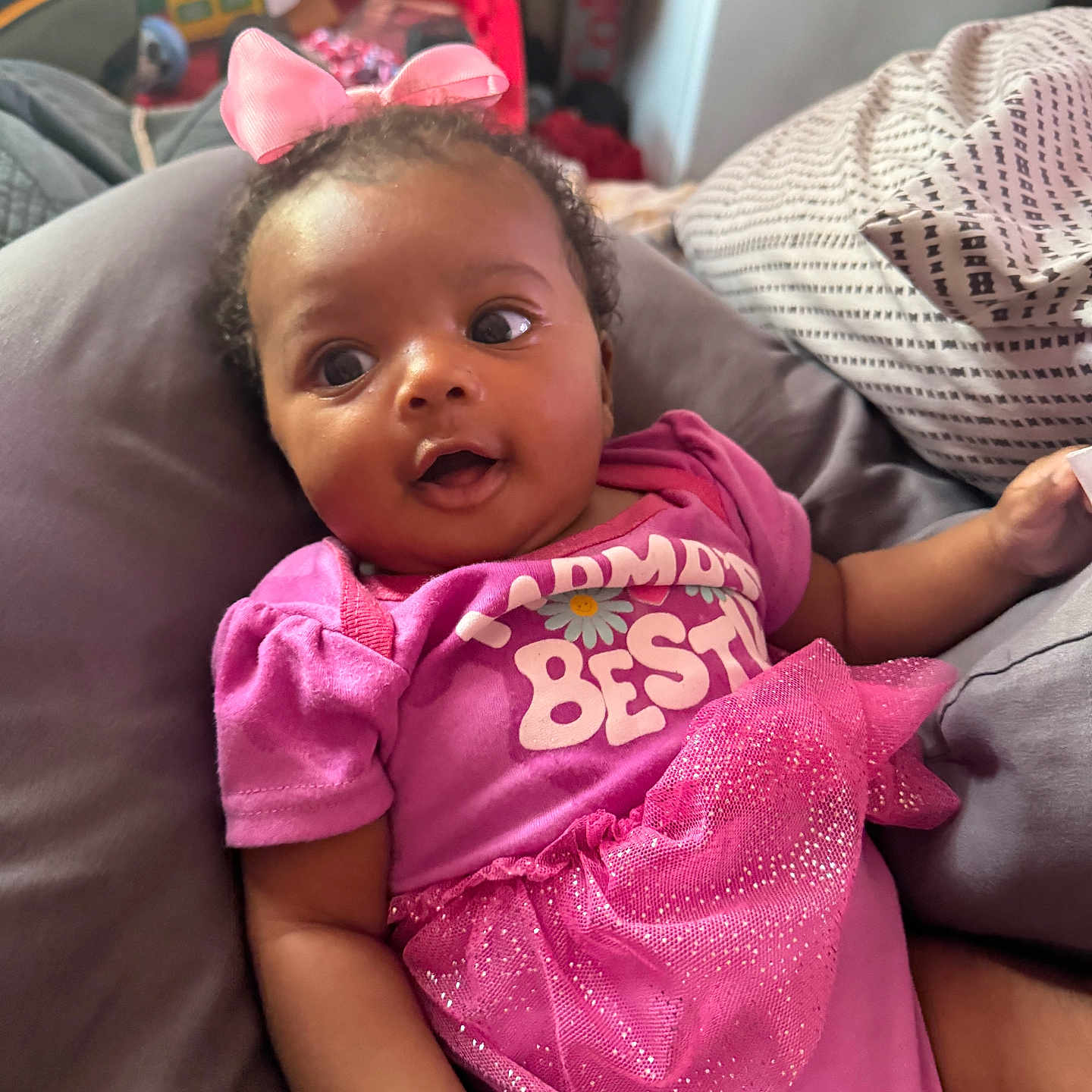 Egypt Ivy Elizabeth is registered to the contest to win money with this photo: baby, child, infant, portrait, indoors, sitting, lying_down, pink_outfit, pink_bow, dress, smiling, wide_eyes, curly_hair, couch, cushions, blanket, toy_storage, home, close_up, fashion