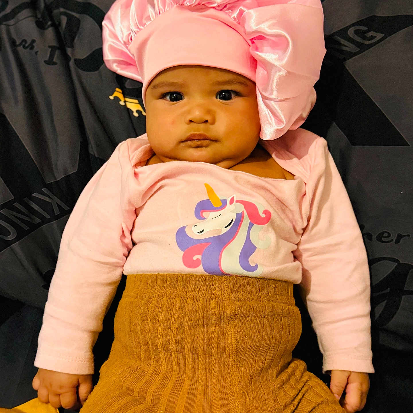 Toahina a rejoint le concours — aidez-le/la à gagner de superbes lots ! baby, child, clothing, cushion, cute, fabric, face, hand, headwear, indoors, infant, knitwear, mustard_pants, person, pink_bonnet, portrait, seated, serious_expression, soft_texture, unicorn_shirt