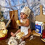 baby, child, chef_hat, apron, flour, dough, bread, baking, kitchen, wooden_table, cutting_boards, bag_of_flour, bowl, crackers, messy, cute, indoors, food, person, smudged_flour