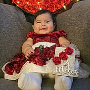 Carla is registered to the contest to win money with this photo: baby, infant, smiling, red_dress, white_tights, couch, flowers, roses, bouquet, lights, celebration, decor, cute, portrait, indoors, holding_sign, happy, child, sitting, cozy