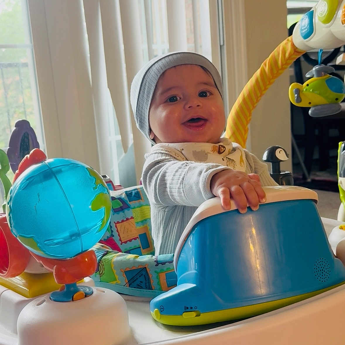 Levi is registered to the contest to win money with this photo: activity_center, baby, beanie, bib, child, clothing, colorful, face, furniture, hand, happy, indoor, infant, person, play, smiling, toy_globe, toy_helicopter, vertical_blinds, window