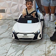 Aliza is registered to the contest to win money with this photo: toddler, child, toy_car, white_car, indoor, marble_floor, geometric_wall, pigtails, curious, steering_wheel, adult_legs, casual_clothing, sneakers, floor_tiles, modern_interior, small_car, electric_car_toy, person, vehicle, cute