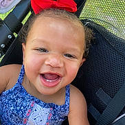Akylah is registered to the contest to win money with this photo: baby, child, smiling, happy, portrait, stroller, red_bow, dress, blue_clothing, infant, face, teeth, outdoor, seated, harness, carriage, close_up, patterned_clothing, joyful, photography