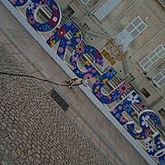 On Duty participe au concours pour gagner de l'argent avec cette photo : architecture, building, cobblestone, colorful_letters, daylight, dog, flower_pattern, leash, no_people, outdoor, pavement, public_space, quiet, shutters, sidewalk, sign, small_dog, street, urban, vintage_style