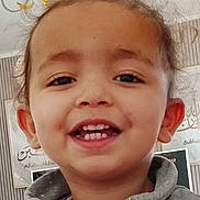 Maïssa a rejoint le concours — aidez-le/la à gagner de superbes lots ! child, toddler, smiling, face, hair, dimples, teeth, clothing, hoodie, indoor, wall_art, calligraphy, portrait, happy, person, topknot, head, expression, curly_hair, background