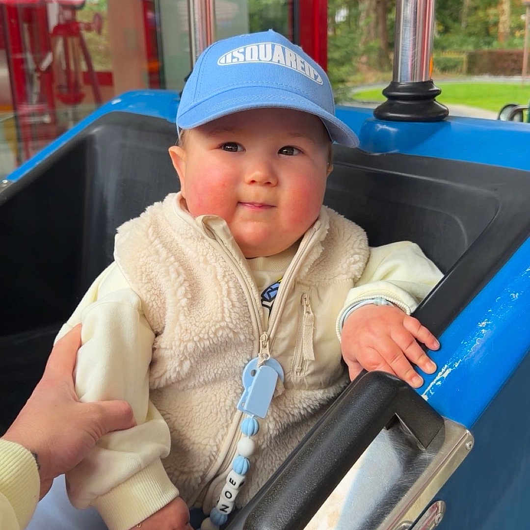 Noe participe au concours pour gagner de l'argent avec cette photo : toddler, child, blue_cap, fleece_vest, amusement_ride, indoor, hand, support, seat, handle, window, greenery, casual_clothing, smiling, face, person, toy_car, white_shoes, play_area, cute