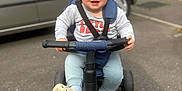 Alfie is registered to the contest to win money with this photo: blond_hair, brick_wall, car, casual, child, clothing, expression, handlebars, outdoor, pavement, person, play, seatbelt, sidewalk, sneakers, toddler, toy, transport, tricycle, vehicle