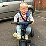 blond_hair, brick_wall, car, casual, child, clothing, expression, handlebars, outdoor, pavement, person, play, seatbelt, sidewalk, sneakers, toddler, toy, transport, tricycle, vehicle