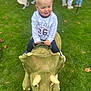Alfie is registered to the contest to win money with this photo: balloon, blonde_hair, blue_pants, blurred_background, casual_clothing, child, dinosaur, event, fun, grass, green, handle, outdoor, park, person, play, toddler, toy, triceratops, white_shirt